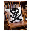 A square crocheted throw pillow with a black and white skull and crossbones design, resting on a wooden spindle-back chair in front of a bookshelf.