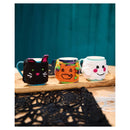 Three crocheted mug cozies shaped like a black cat, a pumpkin, and a ghost, displayed on a wooden tray with blue ceramic mugs against a teal background.