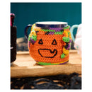A crocheted orange pumpkin-shaped mug cozy with a black jack-o'-lantern face and purple and green trim, wrapped around a dark ceramic mug on a wooden surface.