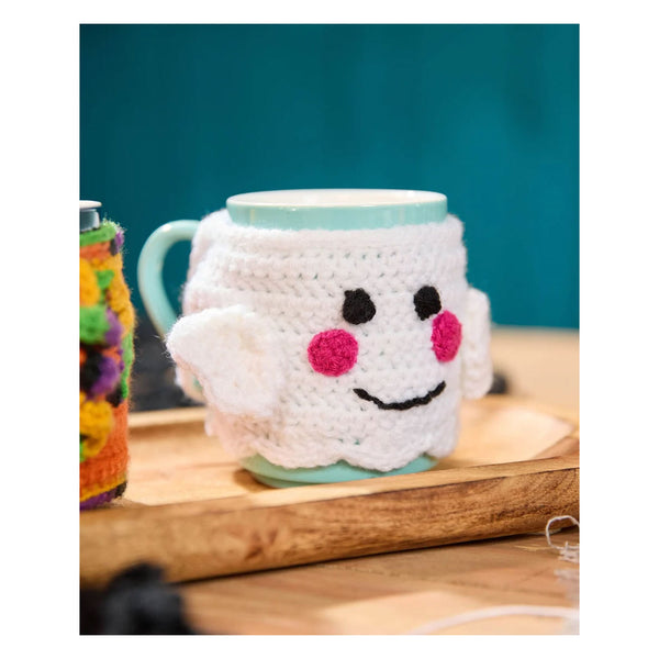 A white crocheted ghost-shaped mug cozy with embroidered black eyes and mouth and pink cheeks, wrapped around a light blue ceramic mug on a wooden tray.