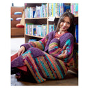 A woman sits on the floor in front of a bookshelf, wearing a large, cozy hand-crocheted cardigan with vibrant multicolor stripes including purple, pink, orange, green, and blue.