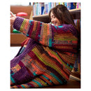 A woman smiling while wearing a large, colorful hand-crocheted blanket or cardigan in a library setting with bookshelves in the background.