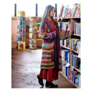 A woman stands in a bookshop aisle wearing a long, colorful hand-crocheted cardigan over a rust-red dress. She is positioned next to a wooden bookshelf filled with books.