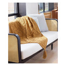A mustard yellow cable-knit throw blanket with a tassel is draped over a modern white and cane daybed in a bright room.