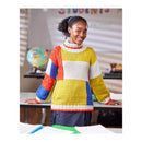 A smiling young woman stands behind a desk in a classroom-like setting, wearing a chunky knit sweater with large color-block panels in yellow, white, red, and blue.