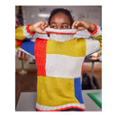 A person wearing a chunky knit turtleneck sweater with large color-blocked panels in yellow, white, red, and blue, holding the collar up to show the fit in an indoor classroom-like setting.