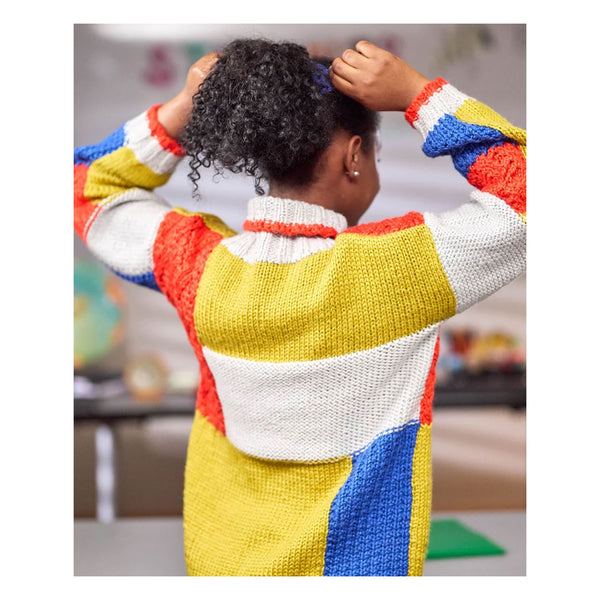 A person shown from behind wearing a chunky knit sweater with large color-blocked panels in yellow, white, red, and blue. The person has curly black hair and is adjusting it with both hands in an indoor setting.