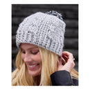 A smiling blonde woman wearing a light gray cable-knit beanie with a black and white pom-pom, shown in a lifestyle portrait against a dark wooden background.