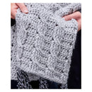 A close-up of a person holding a chunky light gray cable-knit scarf with black and gray fringe tassels.