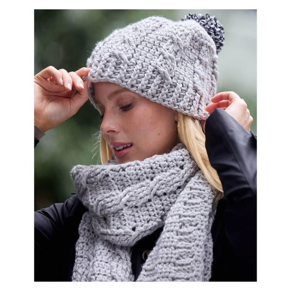 A woman wearing a chunky light gray cable-knit beanie with a black and gray pom-pom and a matching thick knit scarf, photographed outdoors against a blurred green background.