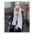 A woman wearing a chunky gray knit beanie, a long gray open-knit scarf, a black coat, and a brown top, standing outdoors in front of a modern building with glass windows.