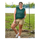 A woman stands outdoors near a lake wearing a sleeveless, open-knit crocheted top in dark teal green and khaki shorts, with white sneakers.