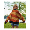 A young woman standing outdoors in a grassy area with trees and a body of water in the background, wearing a chunky knit sweater with wide horizontal stripes in orange, mustard yellow, and dark navy blue.
