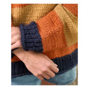 Close-up of a person wearing a chunky knit sweater with wide horizontal stripes in mustard yellow, burnt orange, and dark navy blue, paired with light blue denim shorts.