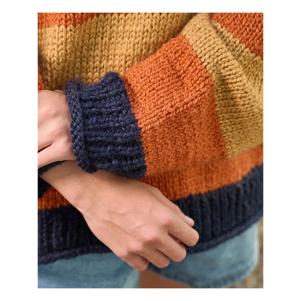 Close-up of a person wearing a chunky knit sweater with wide horizontal stripes in mustard yellow, burnt orange, and dark navy blue, paired with light blue denim shorts.