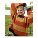 A smiling young woman outdoors wearing a chunky knit sweater with wide horizontal stripes in mustard yellow, rust orange, and dark navy, with her arms raised above her head.