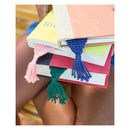 Three colorful crocheted tassel bookmarks in pink, blue, and green are shown draped over a stack of books held by a person.