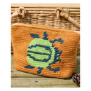 An orange crocheted pouch with a green and blue pixelated character design, displayed on a rustic wooden surface in front of a wicker basket.
