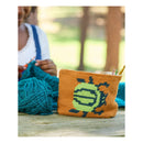 A woven orange pouch with a green and yellow sun design sits on a wooden table in a rustic setting, with a person knitting in the background and a ball of teal yarn in the foreground.