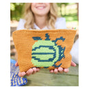 A person outdoors holds a crocheted zip pouch in front of their face. The pouch is mustard yellow with a large green and navy blue graphic design on the front.