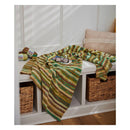A striped knit blanket in green, yellow, brown, and cream is draped over a white bench in a cozy room setting with woven baskets, a throw pillow, and yarn visible in the background.