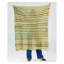 A person holds up a large knit blanket with horizontal stripes in olive green, mustard yellow, and teal, shown against a light gray studio background.