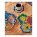A set of handmade crocheted flower-shaped coasters in various colorful patterns, displayed on a wooden table with a white mug filled with coffee.