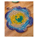 A handmade crocheted flower-shaped potholder with a hanging loop, featuring concentric rings of yellow, teal, blue, beige, and purple yarn on a wooden surface.