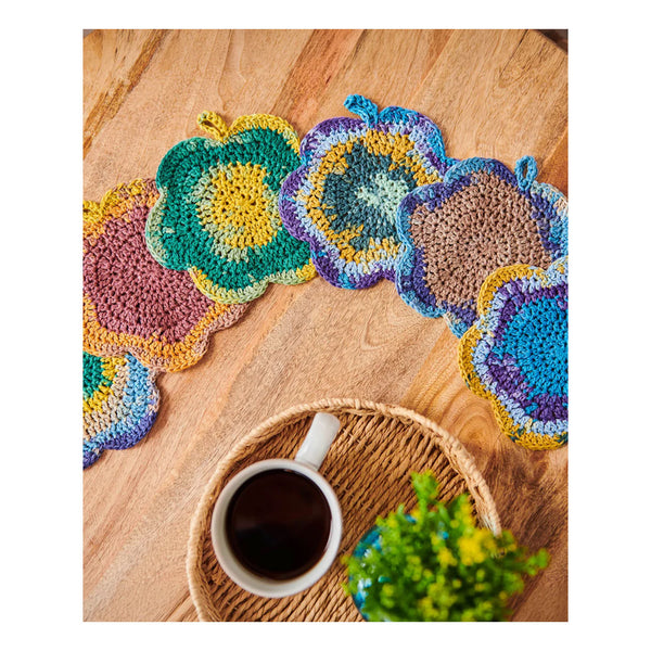 A set of handmade crocheted floral coasters in a multicolor palette of green, blue, purple, yellow, and pink, arranged on a wooden tabletop next to a cup of coffee and a small plant.