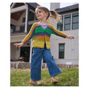 A young girl with a ponytail stands barefoot on a grassy lawn in front of a house, wearing a colorful striped knit cardigan with blue wide-leg jeans.