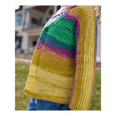A close-up side view of a person wearing a hand-knit cropped cardigan with horizontal stripes in yellow, green, purple, and pink, paired with blue jeans, set against a blurred outdoor background.