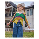 A young girl wearing a colorful striped knit cardigan, blue jeans, and a purple top stands outdoors in front of a house.