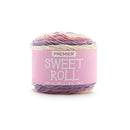 A skein of Premier Sweet Roll yarn with a pink paper label, featuring variegated colors of pink, purple, and cream, set against a plain white background.