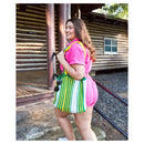 A woman wearing a pink outfit stands in front of a rustic log cabin, carrying a large woven tote bag with green, yellow, and white vertical stripes.