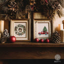 Two framed Christmas needlework or cross-stitch style art prints displayed on a wooden mantel, surrounded by pinecones, red ornaments, candles, and holiday greenery. The left print shows a wreath with a bell, and the right print shows a red pickup truck carrying a Christmas tree.