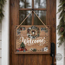 A rustic wooden 'Welcome' door sign hanging on a dark wood front door. The sign features a weathered wood texture, white script lettering, a wreath with a small bell, a red Christmas truck carrying a tree, and holiday floral accents.
