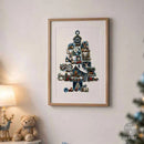 A framed Christmas art print featuring a detailed illustration of a snow-covered tree made of cozy houses, ornaments, and festive details, hung on a light beige wall in a softly lit room with a lamp and teddy bear nearby.