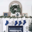 A white fireplace mantel decorated for Christmas, with five stockings hanging below: four navy blue and one light gray. The mantel is garlanded with green pine branches, decorated with small framed pictures, a green wreath in an arched mirror, and a white Christmas tree.