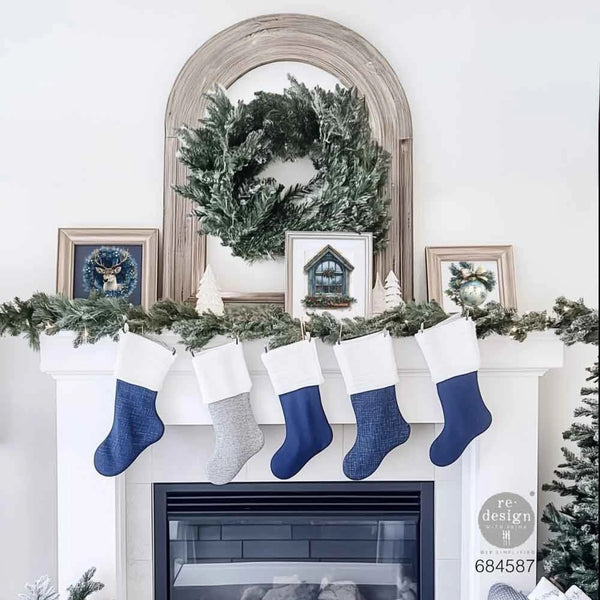 A white fireplace mantel decorated for Christmas, with five stockings hanging below: four navy blue and one light gray. The mantel is garlanded with green pine branches, decorated with small framed pictures, a green wreath in an arched mirror, and a white Christmas tree.