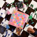 A colorful square art piece with a pink border and multicolored checkered squares sits on a black-and-white checkered floor, surrounded by Rit paint bottles, markers, and green paint tubes. Two hands are visible holding markers over the artwork, making this a lifestyle shot.