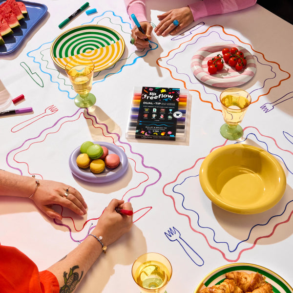 A tabletop scene with multiple people's hands visible, drawing colorful wavy lines on a white surface using markers. The image includes plates with strawberries, macarons, watermelon, yellow bowls, glasses, and a boxed set of markers labeled 'big freeflow'.