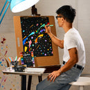A man wearing glasses and a white t-shirt sits at an easel, drawing on a black canvas with colorful paint. In the foreground, a small table holds a box of 'Big Flow' markers and several marker pens.