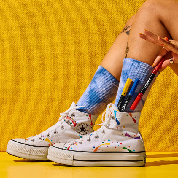 White Converse high-top sneakers with colorful paint splatter details, worn with blue tie-dye crew socks. A hand is holding red, blue, yellow, and black markers that are sticking out of the socks, set against a bright yellow background.