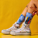 White Converse high-top sneakers with colorful paint splatter details, worn with blue tie-dye crew socks. A hand is holding red, blue, yellow, and black markers that are sticking out of the socks, set against a bright yellow background.
