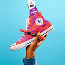A hand holds a Converse All Star high-top sneaker with a purple and pink paint-splatter pattern, with a tube of Rit Purple Paint sticking out of the collar, against a bright blue background.