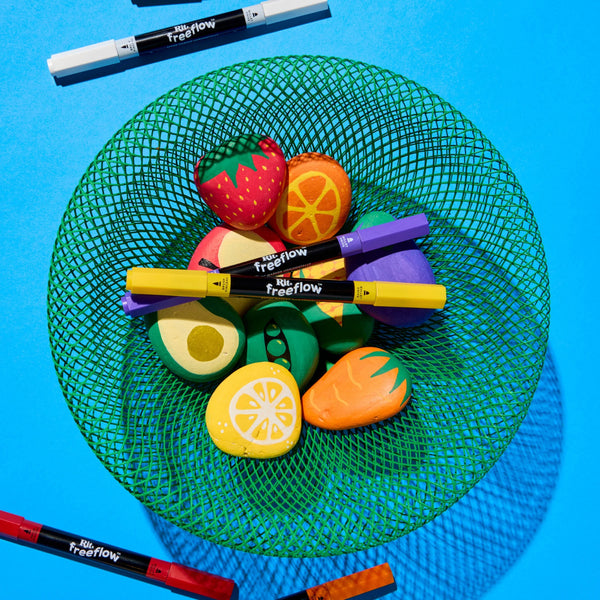 A top-down flat lay of colorful fruit-shaped erasers in a green mesh bowl, with several black Treflows markers placed around and across the bowl on a bright blue background.