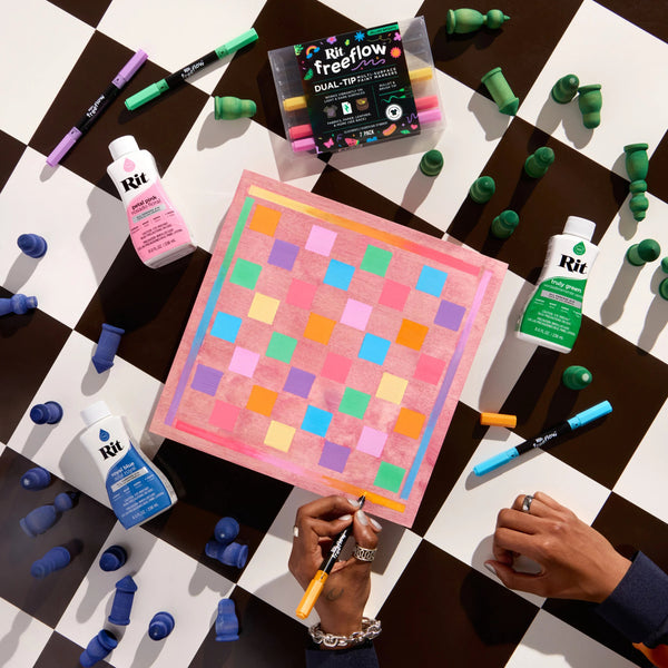 A colorful square art piece with a pink border and multicolored checkered squares sits on a black-and-white checkered floor, surrounded by Rit paint bottles, markers, and green paint tubes. Two hands are visible holding markers over the artwork, making this a lifestyle shot.