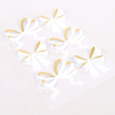 Five white paper bow stickers with gold accents and small gold polka dots, arranged on a clear sheet over a light beige background.