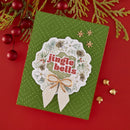 A handmade Christmas greeting card with a green textured background and a central white scalloped wreath design reading 'jingle bells' in red letters, decorated with pinecones, evergreen branches, and a cream pleated bow. The card is photographed on a red background with holiday ornaments and pine branches around it.