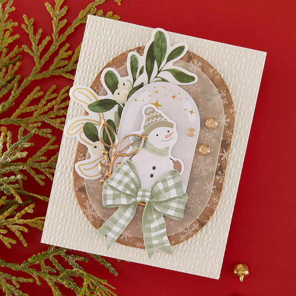 A handmade Christmas greeting card featuring a die-cut snowman, green leaves, and a green-and-white gingham bow, displayed on a textured white cardstock over a gold oval layer against a red background with gold pine branches.